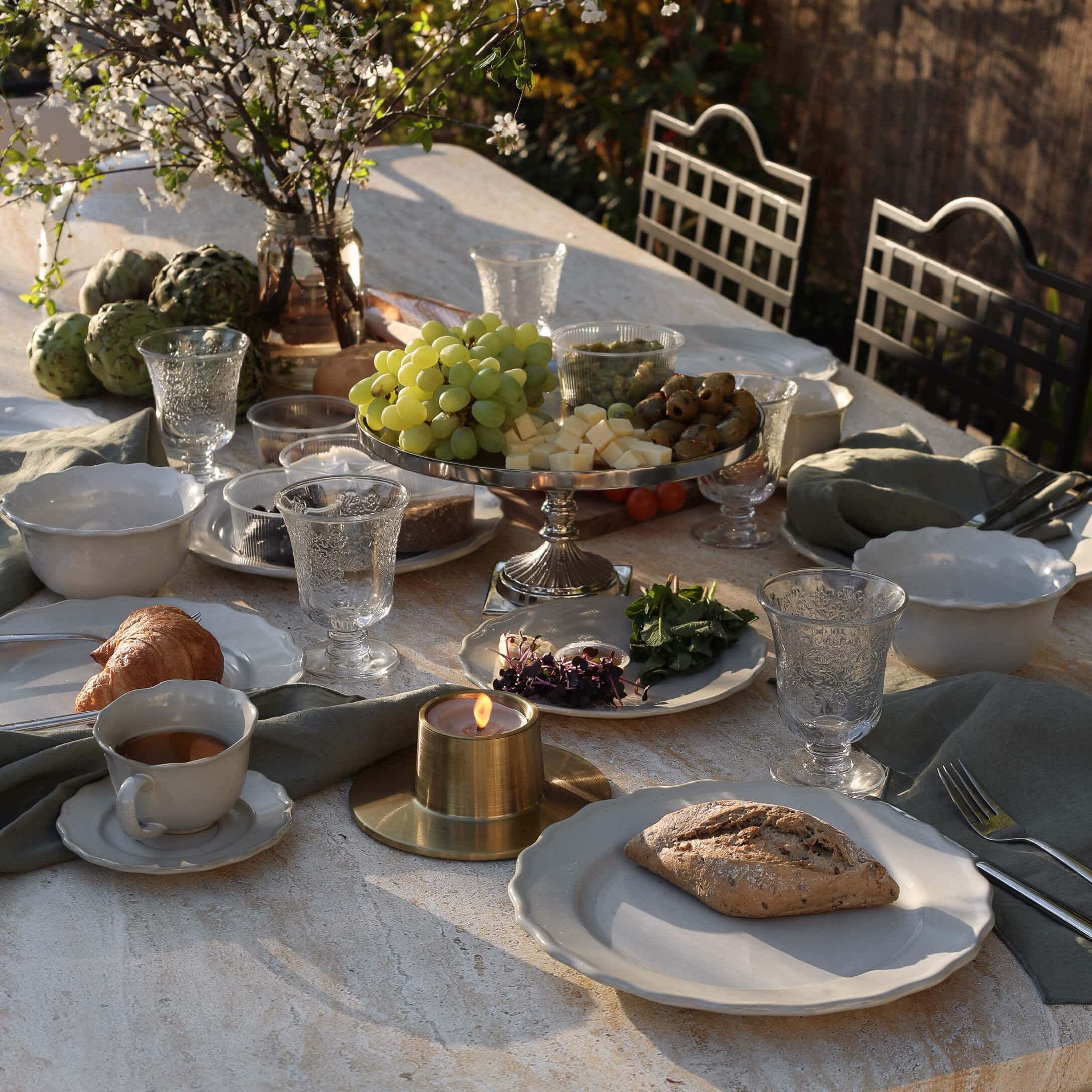 Mesa de piedra con vajilla blanca, vela y bandeja con uvas, queso y aceitunas al aire libre