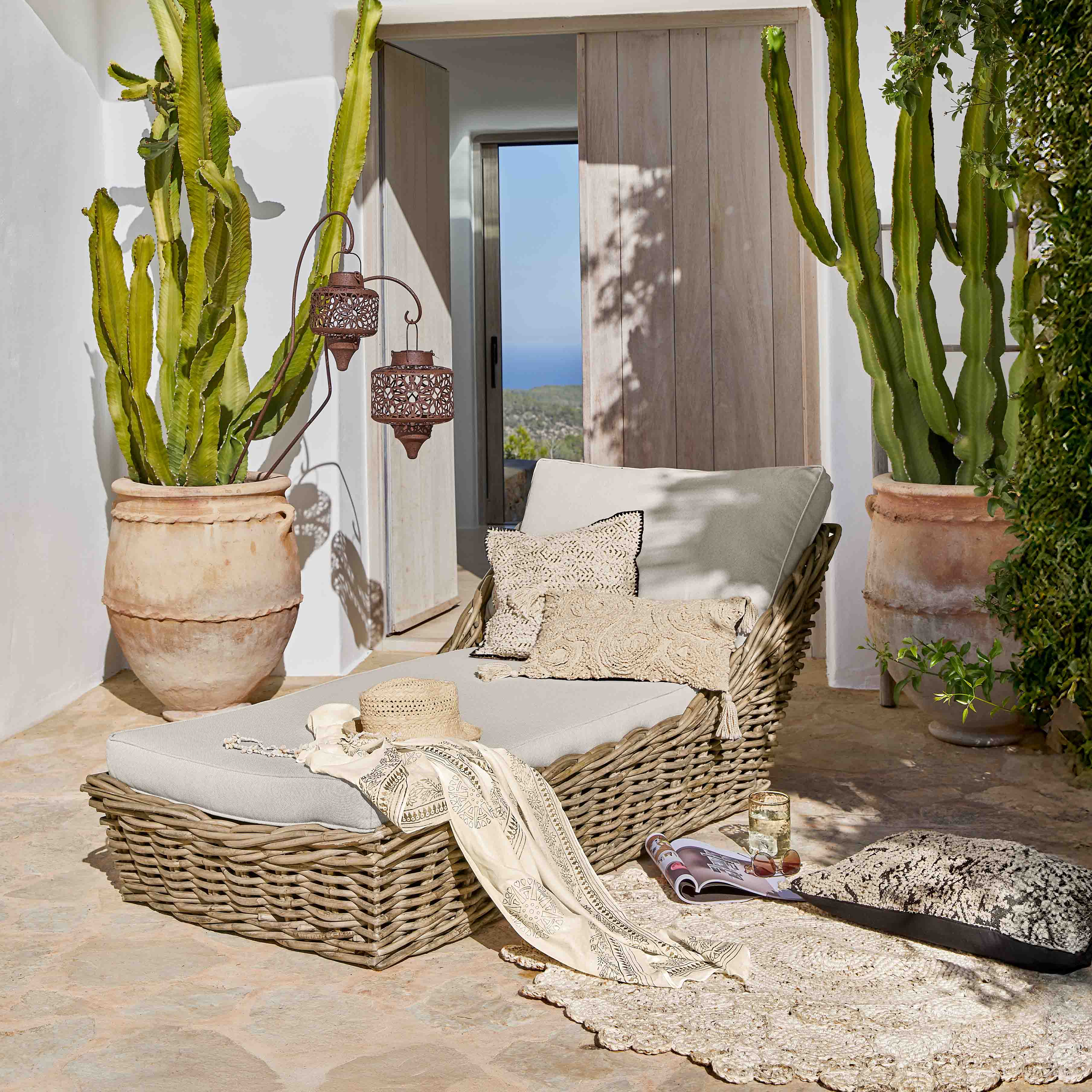 Tumbona de mimbre con cojines gris claro en terraza con grandes cactus en macetas