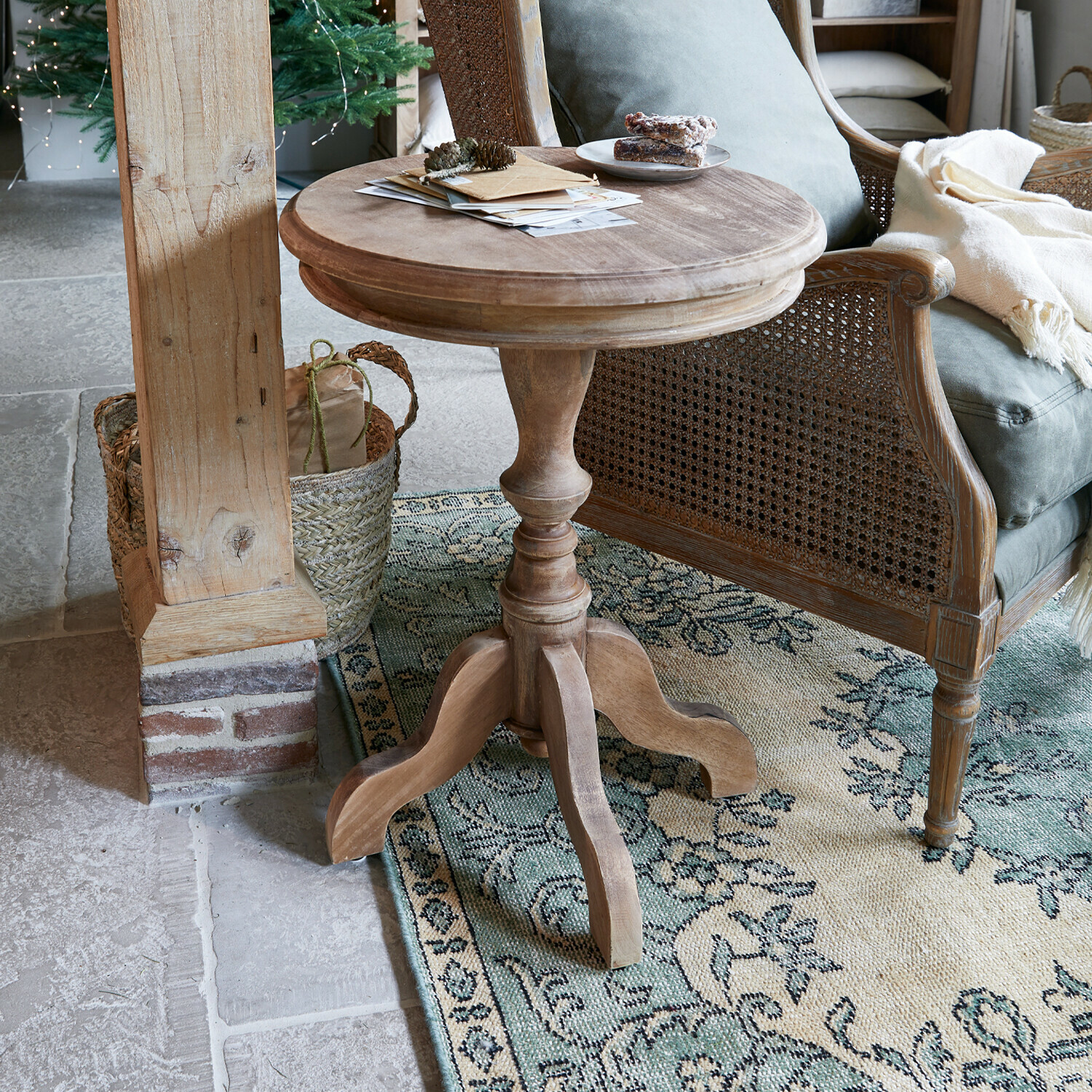 Mesa auxiliar Mengomeyen Mesa redonda de madera natural junto a un sillón de mimbre sobre una alfombra con diseño