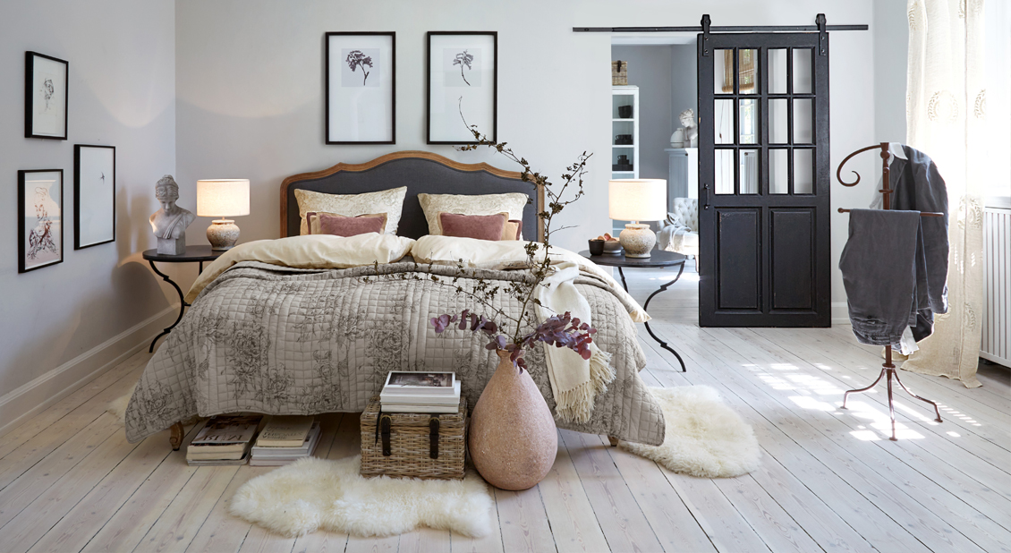 Dormitorio acogedor con cama en ropa de cama gris-beige, mesitas negras y jarrón decorativo con ramas