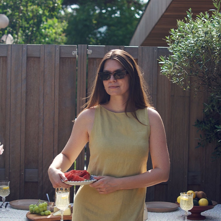 Mujer con vestido amarillo de lino sosteniendo un plato con sandía frente a una cerca de madera y mesa con frutas