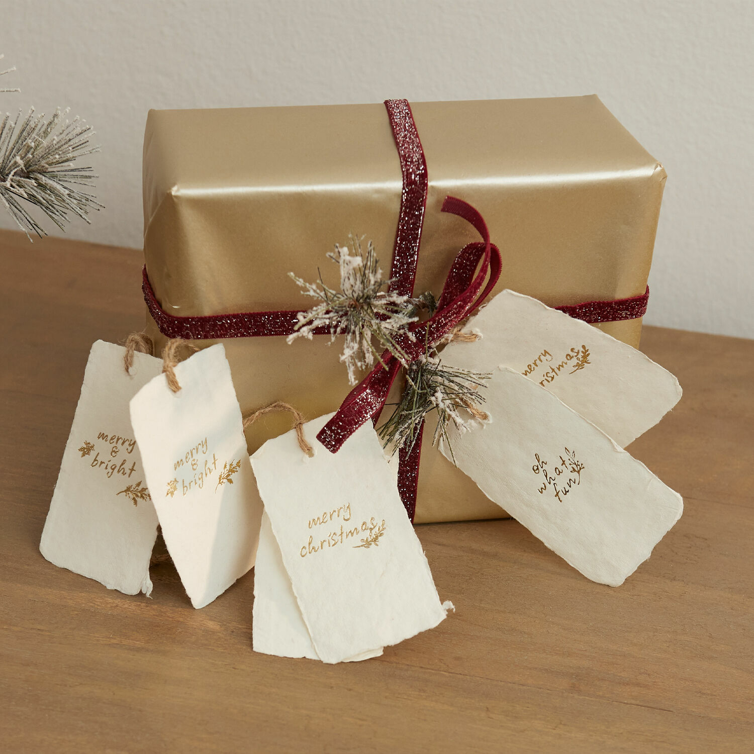 Set de 6 etiquetas Aamira Regalo dorado con cinta burdeos y etiquetas de papel blancas navideñas sobre mesa de madera