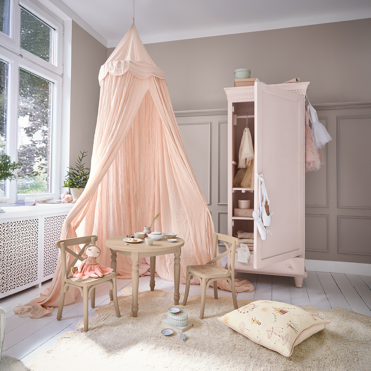 Acogedora habitación infantil con dosel rosa, mesa y sillas de madera, armario rosa abierto con ropa