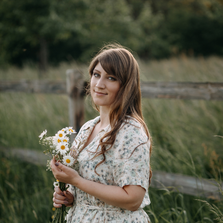 Mujer con vestido floral sosteniendo ramo de margaritas frente a campo verde y cerca de madera