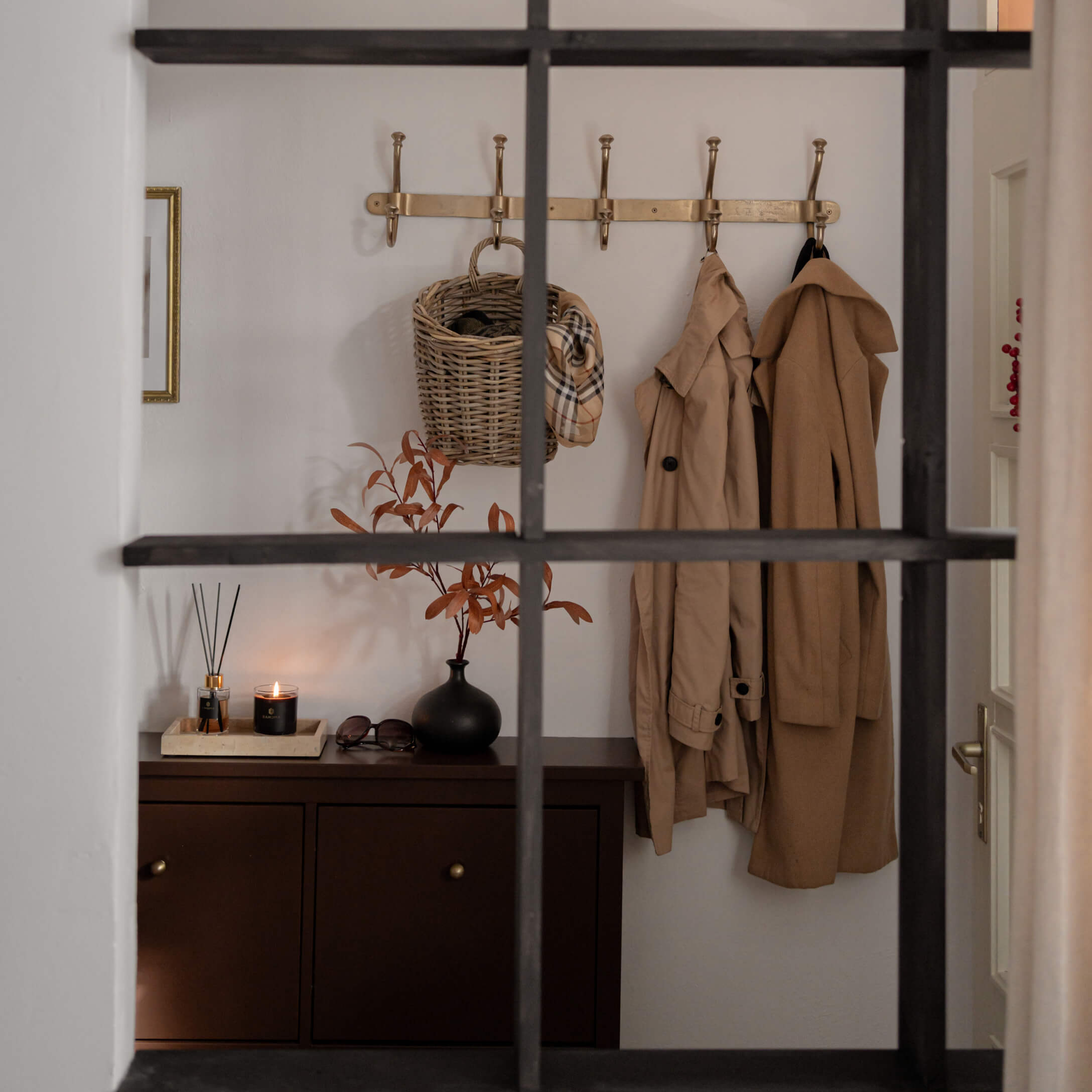 Recibidor con mueble marrón, abrigos beige y cesta de mimbre en perchero dorado