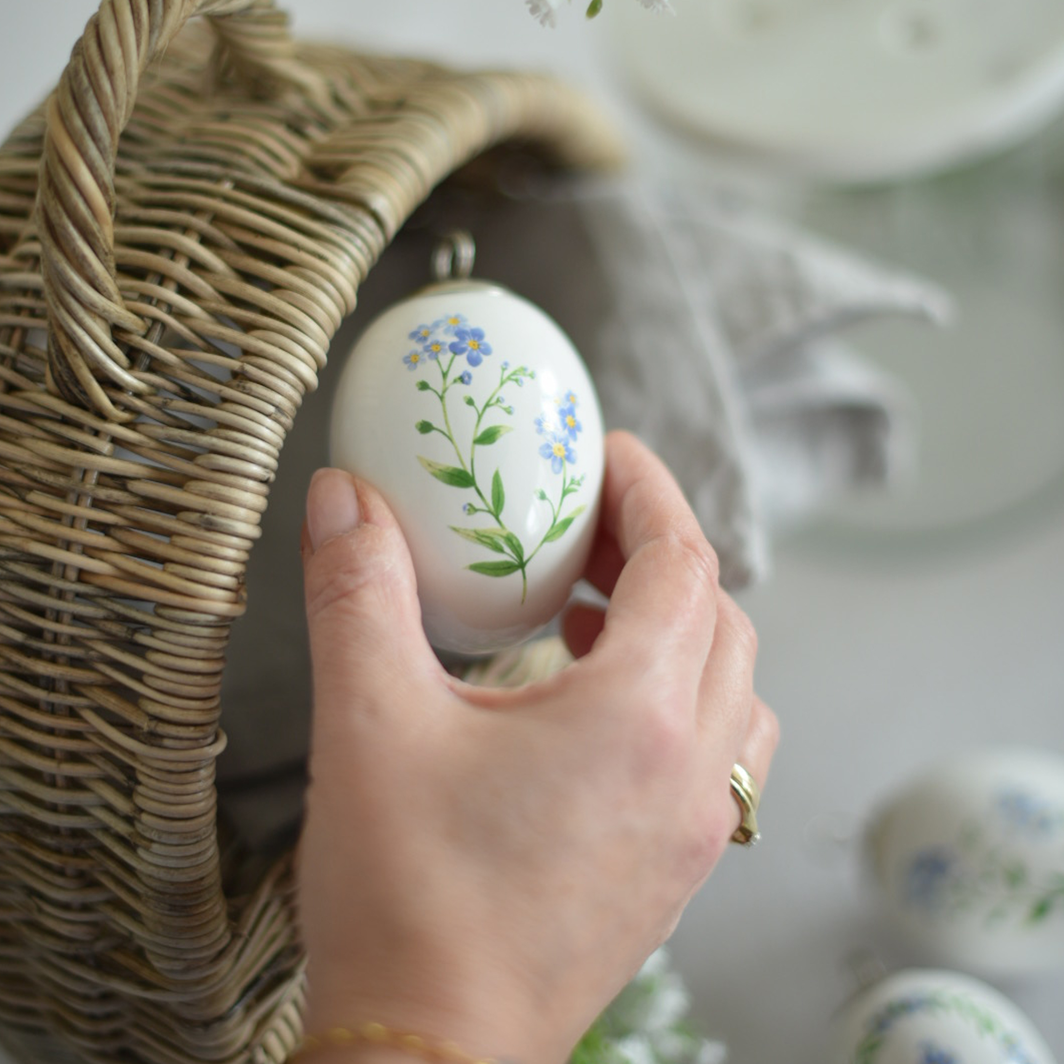 Mano sosteniendo adorno de Pascua de cerámica blanca con flores azules junto a cesta de mimbre