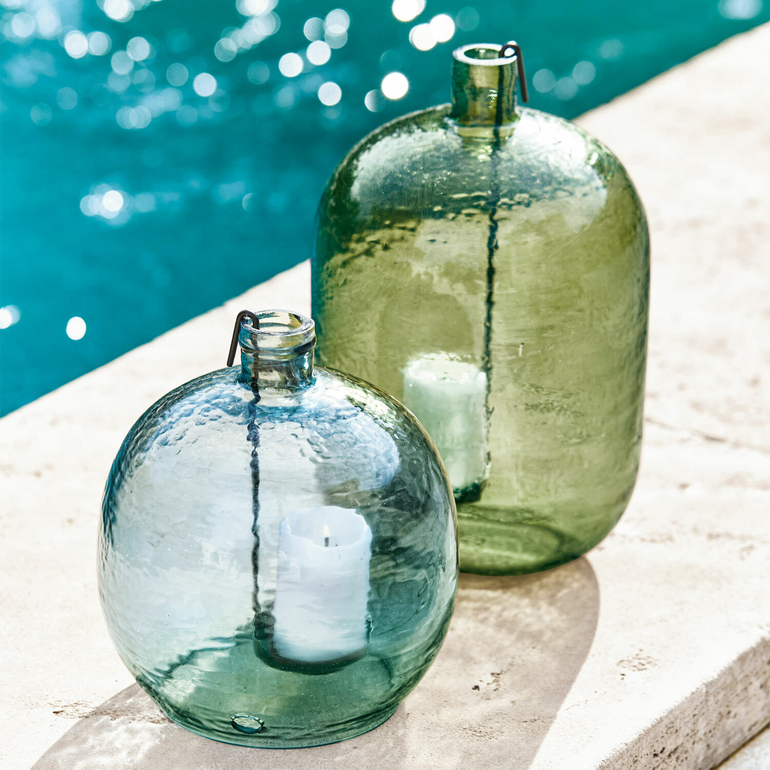 Set de 2 portavelas Michel Faroles de vidrio verde y azul con velas blancas en terraza junto a la piscina