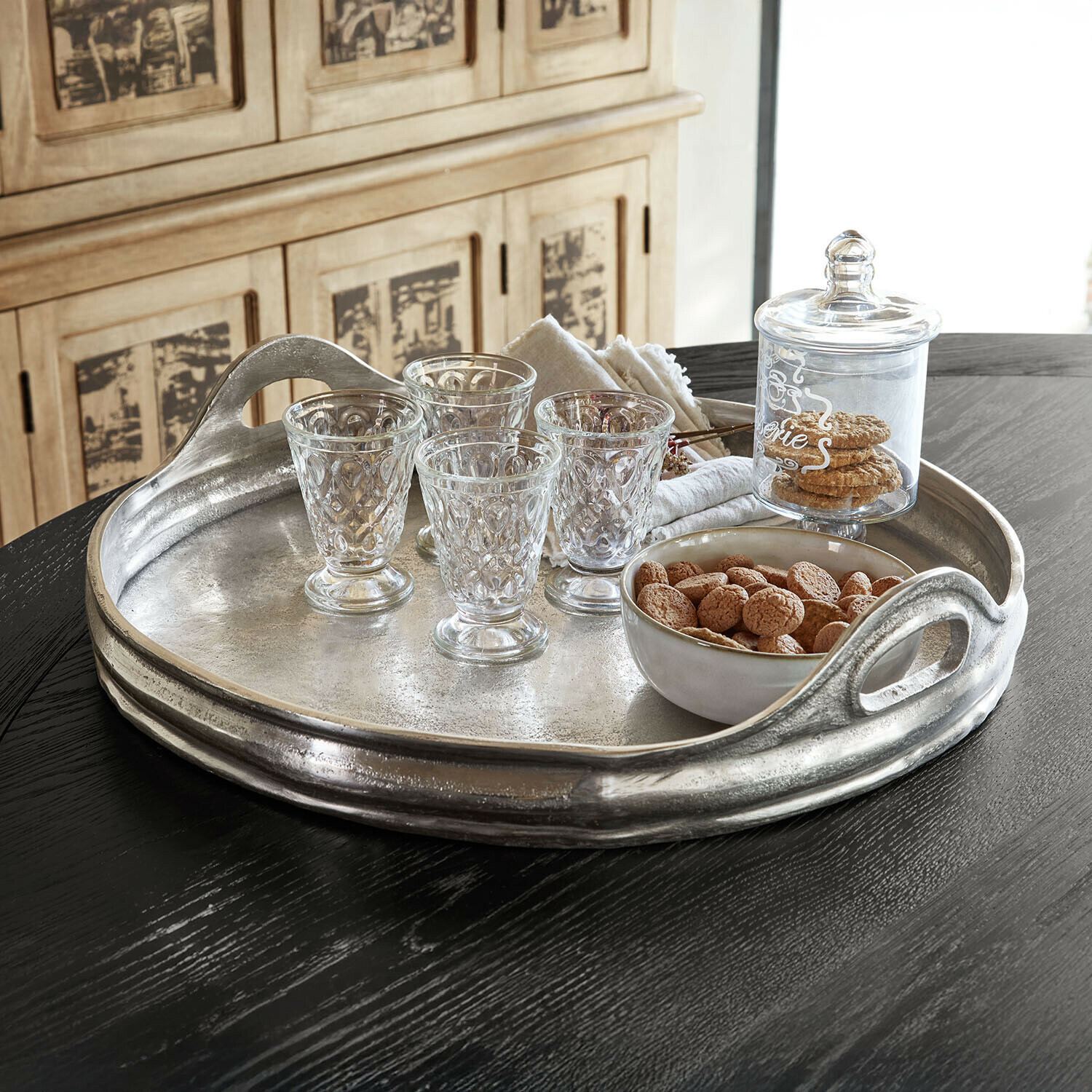 Bandeja Téloriane Bandeja metálica con asas, vasos de cristal y cuenco con galletas sobre mesa negra para servicio elegante