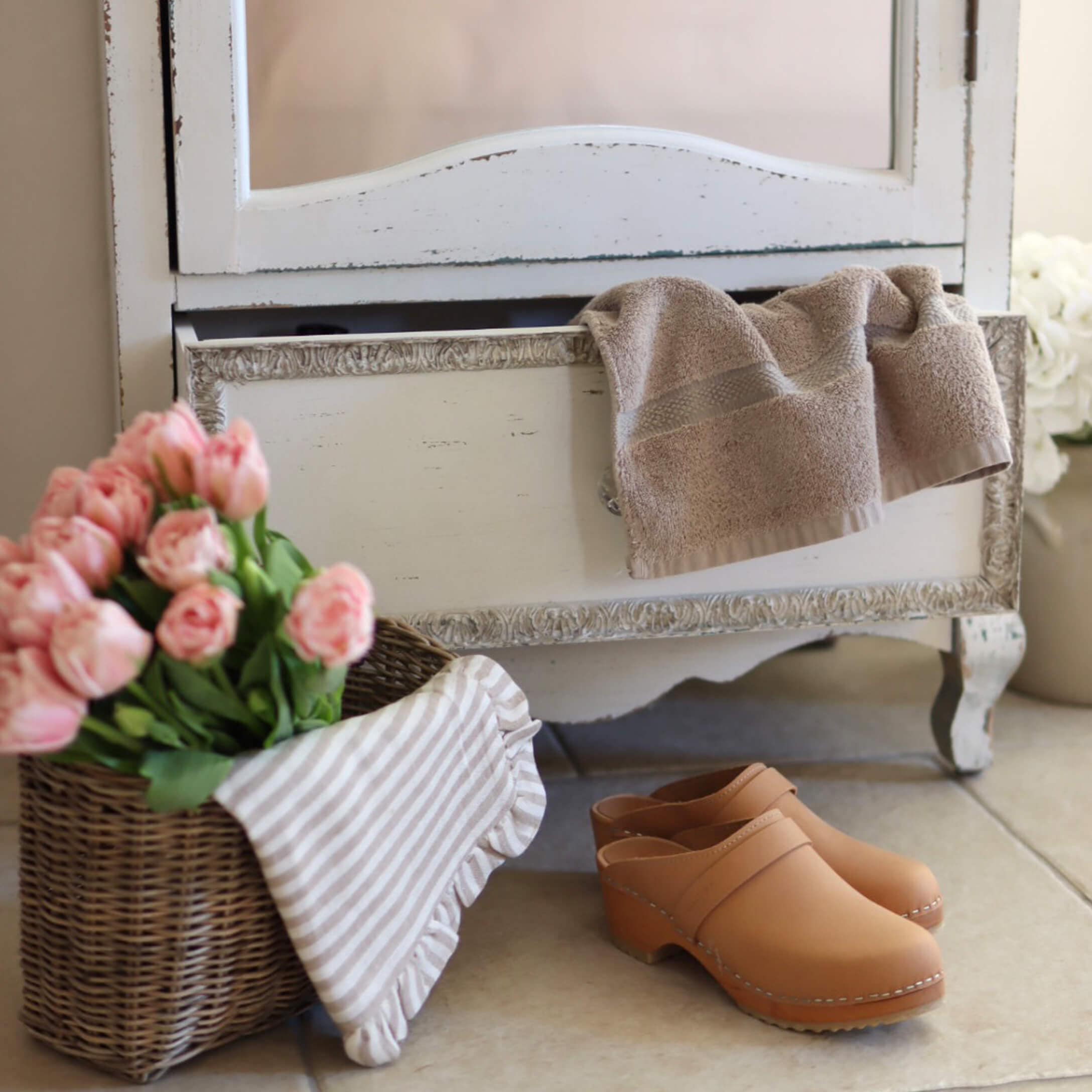 Zuecos de cuero beige en el suelo junto a cesta de mimbre con flores rosas y toalla