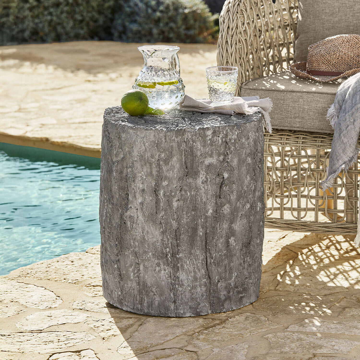 Mesa auxiliar Falmont Mesa auxiliar gris de hormigón con textura de madera junto a sillón de mimbre junto a la piscina