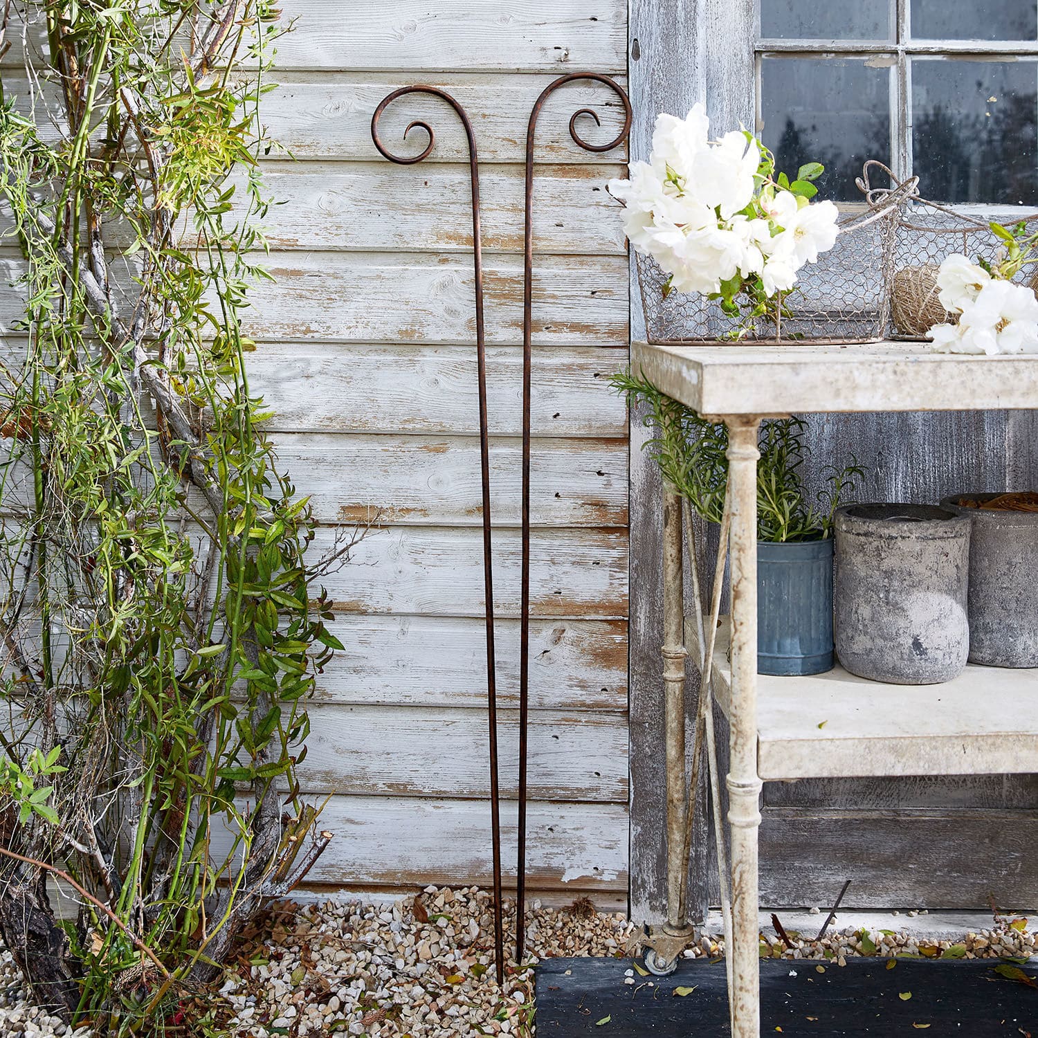 Set de 2 estacas de jardín decorativas Dorhal Soporte rústico de metal oxidado para plantas junto a pared blanca de madera en jardín