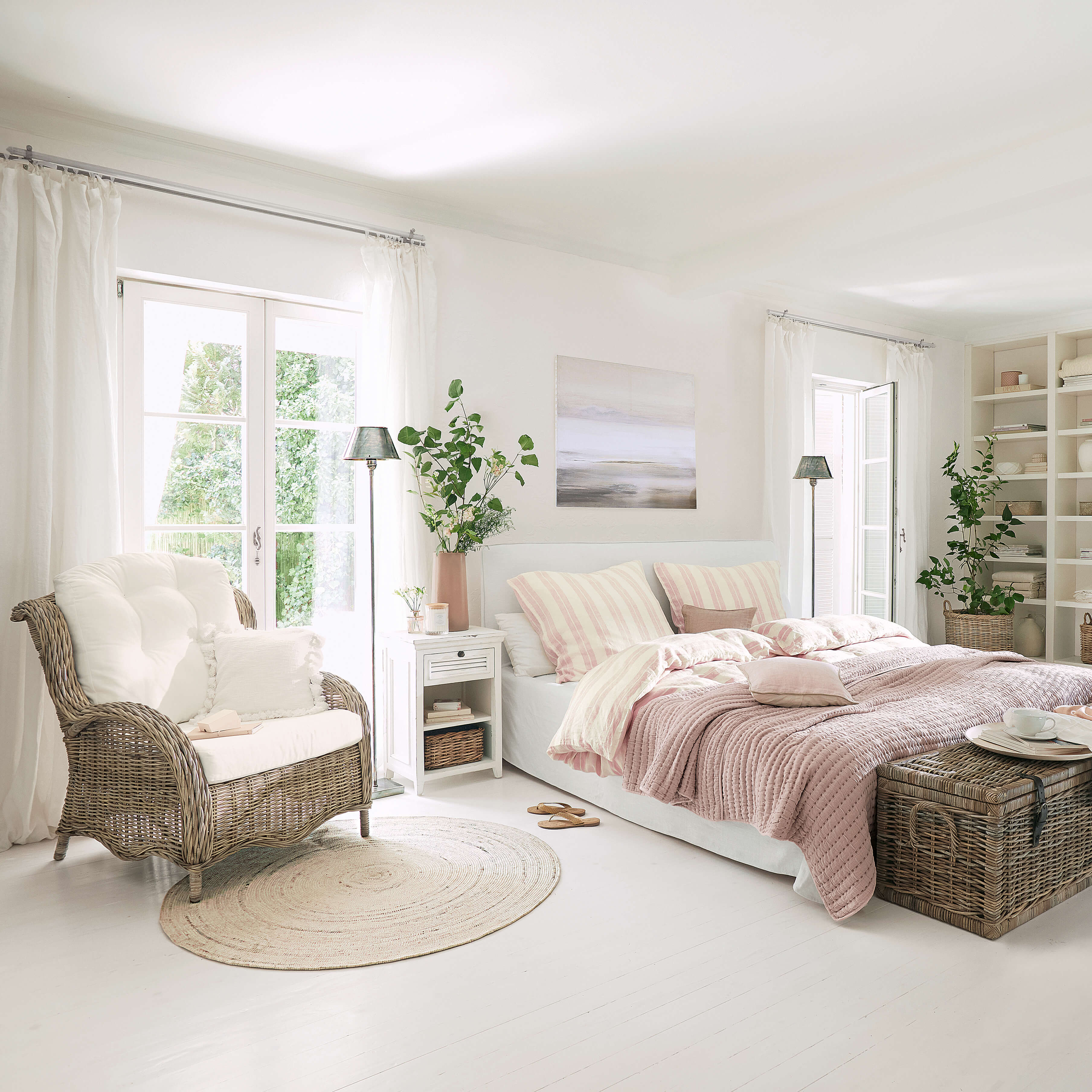 Dormitorio luminoso con cama blanca, ropa de cama rosa y sillón de mimbre sobre suelo claro