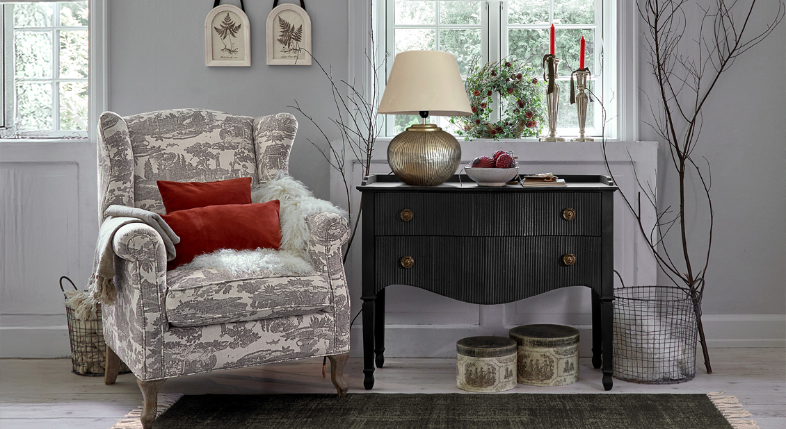 Sillón Toile de Jouy gris y blanco con cojines rojos y cómoda negra en sala luminosa