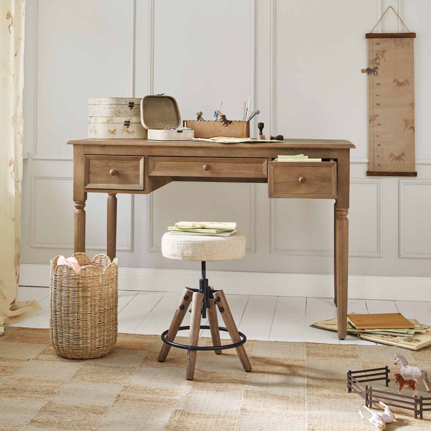 Escritorio de madera con tres cajones y taburete beige frente a pared clara con decoración de caballos